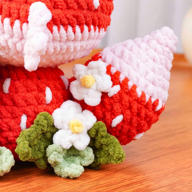 May include: Close-up of a handmade crochet strawberry. The strawberry is red with white seed details and a light pink top. White flowers with yellow centers and green leaves are attached to the strawberry.