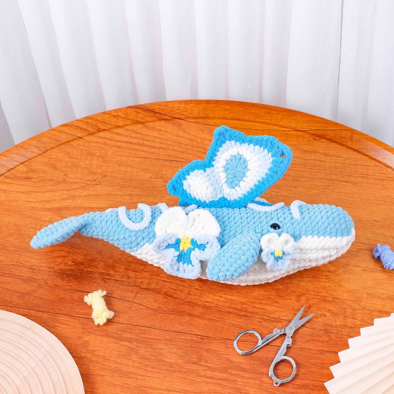 May include: A handmade crochet whale in shades of blue and white, decorated with flower and butterfly details. The whale is on a wooden surface. Small scissors are also visible.