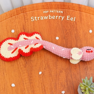 May include: A crocheted Strawberry Eel toy in pink, red, and cream colors, lying on a wooden surface. The toy has a guitar-like shape with the text "Strawberry Eel" above it. The toy is next to two small potted plants.