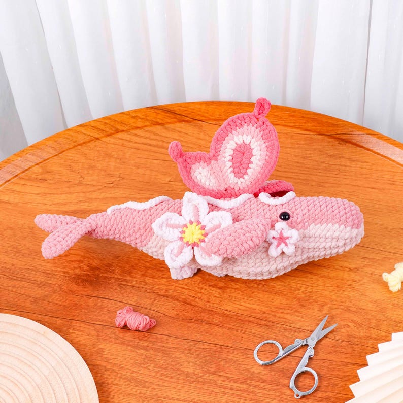 May include: A handmade crochet whale in shades of pink and white, decorated with a pink butterfly and flower details. The whale has a small eye and a tail. The craft project is on a wooden surface.
