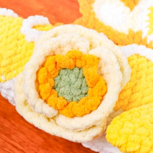 May include: Close-up of a handmade crochet flower. It has a cream-coloured outer layer, a bright orange middle, and a green centre. Other crochet pieces in yellow and white surround the flower, creating a textured, floral design.
