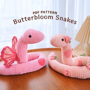 May include: Two crocheted pink snake plushies with flower accents. The snake on the left has a butterfly wing design. The text "PDF PATTERN Butterbloom Snakes" is above the plushies.