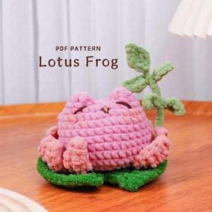 May include: A handmade crochet frog in a lotus flower design. The frog is pink with a smiling face and sits on a green lily pad. The text "PDF PATTERN Lotus Frog" is visible.