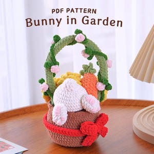Può includere: Un coniglietto all'uncinetto in un cestino marrone con una vite verde e fiori rosa. Il cestino ha un nastro rosso. Il titolo "PDF PATTERN Bunny in Garden" è in cima all'immagine.