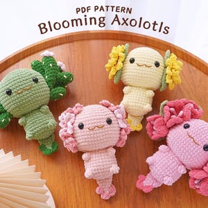 May include: Four crocheted axolotl plushies in different colors. The axolotls are wearing flower crowns and have embroidered eyes and mouths. The axolotls are sitting on a wooden surface. The text "PDF PATTERN Blooming Axolotls" is at the top of the image.