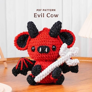 Op de afbeelding: Een handgemaakte rode en zwarte Evil Cow gehaakte knuffel met hoorns, vleugels en een drietand. De tekst "PDF PATTERN Evil Cow" is zichtbaar. De koe zit op een houten oppervlak.