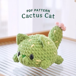 May include: A crocheted cactus cat plush toy with a pink flower on top. The toy is green and has a cute face with black eyes and a yellow nose.