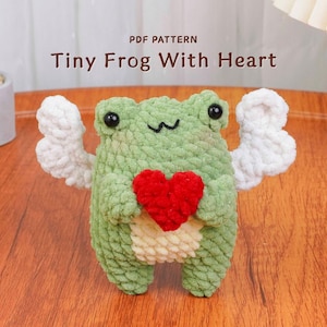 May include: A handmade green crochet frog with white wings and a red heart. The frog has black eyes and a smiling mouth. The text "PDF PATTERN Tiny Frog With Heart" is displayed above the frog.
