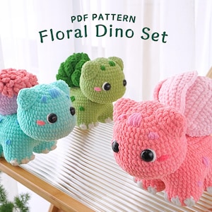 May include: Three crocheted dinosaur plushies in a set. The dinosaurs are green, blue, and pink. The green dinosaur has a leafy green head. The blue dinosaur has a pink flower on its back. The pink dinosaur has a pink flower on its head. The text "PDF PATTERN Floral Dino Set" is at the top of the image.