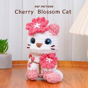 Può includere: Un gatto all'uncinetto fatto a mano in bianco e rosa, adornato con dettagli di fiori di ciliegio. Il gatto ha occhi blu e baffi neri. Il testo "Cherry Blossom Cat" è visualizzato sopra il gatto, indicando un modello PDF.