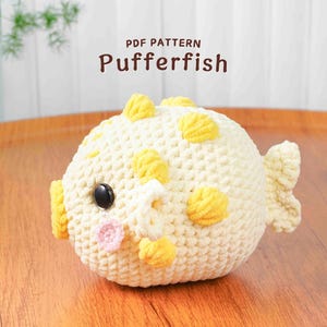 May include: A cream and yellow crocheted pufferfish toy with a black eye and pink details. The handmade plush has yellow spots and fins. The text "PDF PATTERN Pufferfish" is at the top.