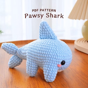 May include: A light blue crocheted shark plush toy with black eyes and a pink mouth. The toy is sitting on a wooden surface. PDF PATTERN Pawsy Shark