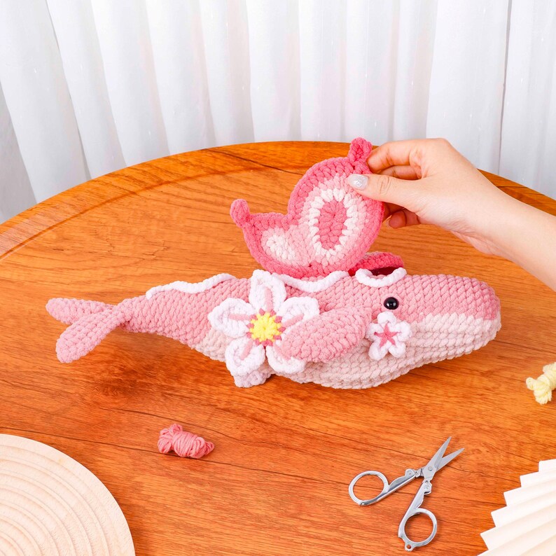 May include: A handmade pink and white crocheted whale with butterfly and flower details. The whale features a small black eye and a tail. A hand holds a pink butterfly. The whale is on a wooden table.