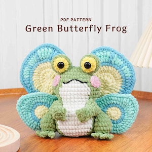 May include: A handmade crochet frog with butterfly wings. The frog is green with a white belly, pink cheeks, and large yellow eyes. The wings are blue and yellow. The text "PDF PATTERN Green Butterfly Frog" is at the top.