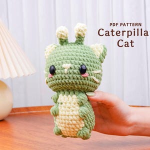 May include: A handmade green and cream-colored crochet Caterpillar Cat plushie. The amigurumi has large black eyes, pink cheeks, and white whiskers. The text "PDF PATTERN Caterpillar Cat" is displayed in brown.