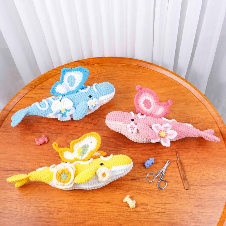May include: Three crocheted whale ornaments in blue, pink, and yellow, each with butterfly wings and floral details. The whales are displayed on a wooden surface, highlighting the handmade quality and bright colours.