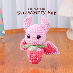 Pode incluir: Um morcego rosa de crochê com corpo e asas de morango. O morcego tem olhos grandes e pretos e a inscrição "PDF PATTERN Strawberry Bat". Sobre uma superfície de madeira.