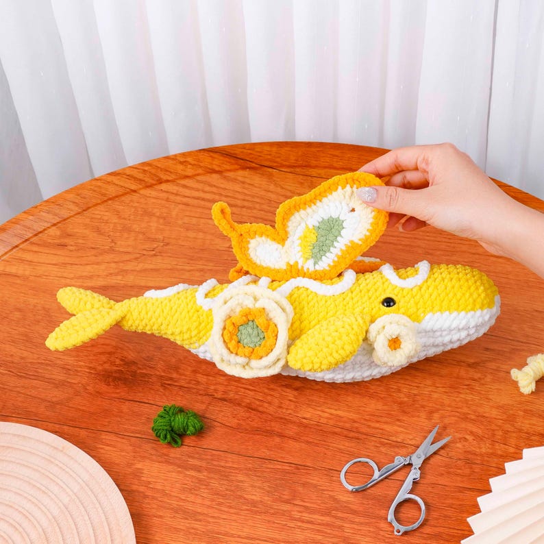May include: A handmade yellow and white crochet whale with a yellow and white butterfly on top. The whale has a flower design on its side. The butterfly is being held by a hand. The whale is on a wooden table.