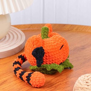 May include: A handmade orange crochet pumpkin with a black patch and a green stem. The pumpkin has a curved, striped tail in orange and black. The pumpkin is a Halloween or fall-themed decorative item.