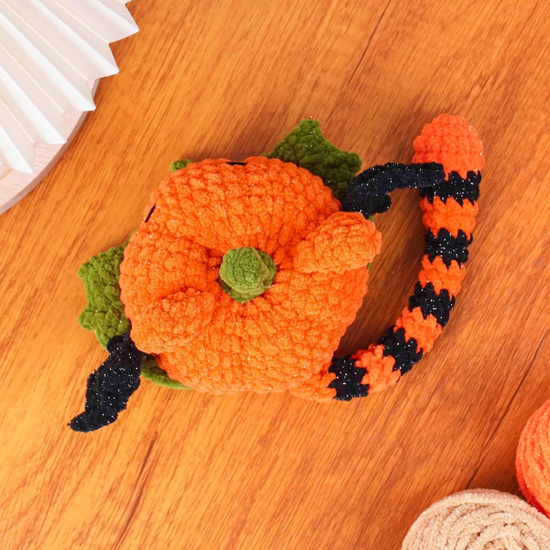 May include: A handmade, orange crochet pumpkin with green leaves and a black and orange striped handle. The pumpkin has a green stem and is likely a Halloween decoration.