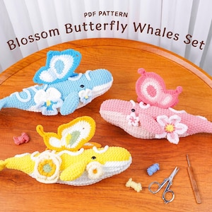 May include: Three crocheted whale toys in blue, pink, and yellow, each with a butterfly wing and flower accents. The text "Blossom Butterfly Whales Set" is visible. Crochet hook and scissors are also present.