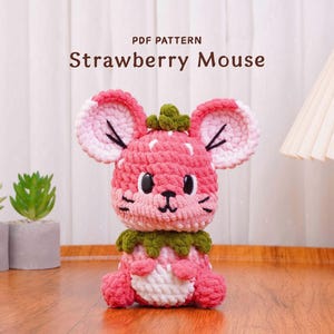 May include: A handmade crochet strawberry mouse in shades of pink, white, and green. The mouse has large ears, a green leafy top, and a white belly. The text "PDF PATTERN Strawberry Mouse" is displayed above the mouse.