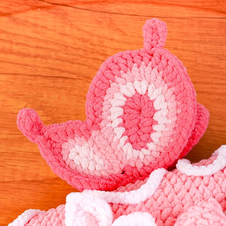 May include: A handmade, pink and white crocheted snail-shaped item. The snail has a spiral shell design with varying shades of pink and white. The body is a solid pink colour. The item is on a wooden surface.
