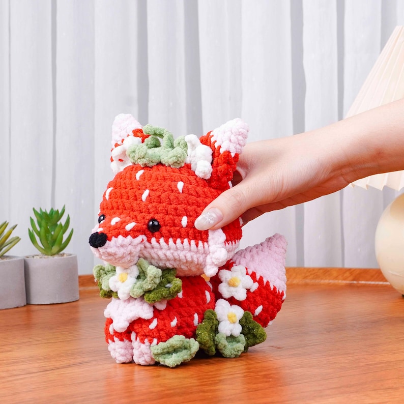 May include: A handmade crochet fox figurine in red, white, and pink. The fox is adorned with green and white floral accents. The fox is being held by a person.