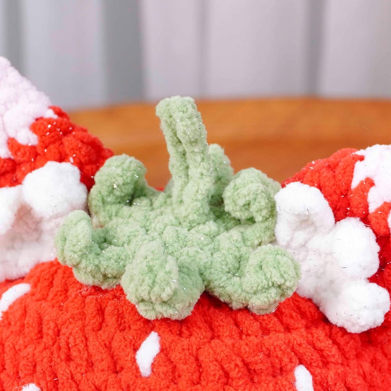 May include: Close-up of a handmade, knitted strawberry. The strawberry is primarily red with white seed details and a green stem. The texture of the yarn is visible, creating a soft, plush appearance. The craft item is likely a decorative piece.