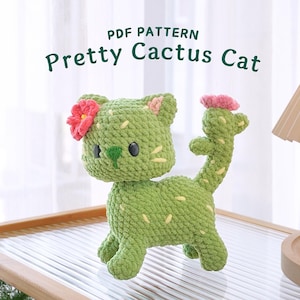 Puede incluir: Patrón PDF para tejer un gato cactus verde con una flor rosa en la cabeza. El gato está sentado en una mesa de madera con una lámpara blanca en el fondo. El texto "PDF PATTERN Pretty Cactus Cat" está en la parte superior de la imagen.
