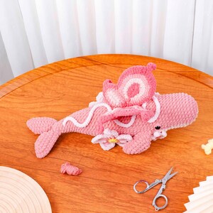 May include: A handmade pink crocheted whale with white detailing, a flower, and a decorative wing. The whale is on a wooden surface. Small scissors and a ball of pink yarn are also visible.