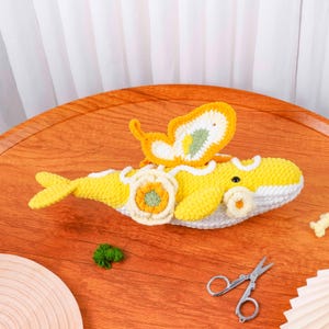 May include: A crocheted whale toy in yellow and white, with a yellow and white butterfly attached. The whale features a flower motif. The toy is displayed on a wooden table.