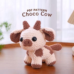 May include: A crocheted brown and white cow amigurumi toy with black eyes. The toy is sitting on a wooden surface. The text "PDF PATTERN Choco Cow" is displayed above the toy.
