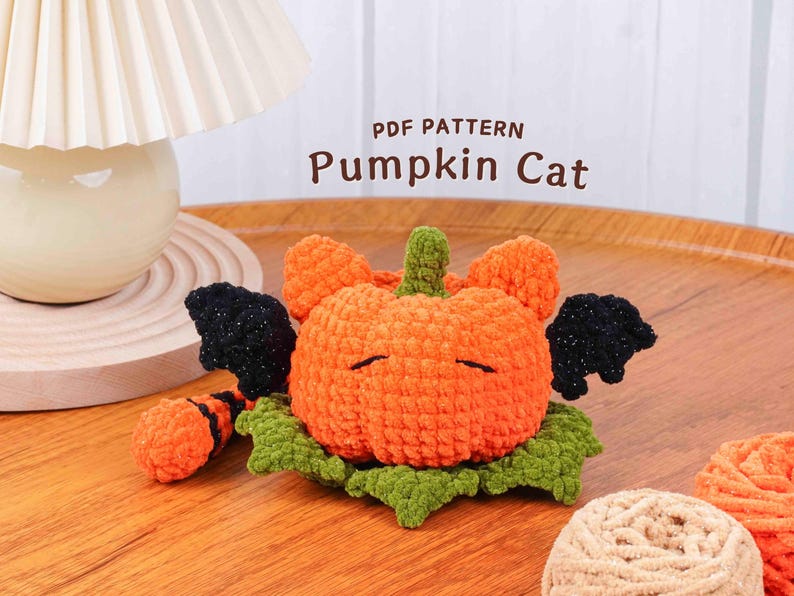 May include: A crocheted pumpkin cat with orange body, green stem and leaves, and black wings. The text "PDF PATTERN Pumpkin Cat" is displayed above the cat. The cat is on a wooden surface.
