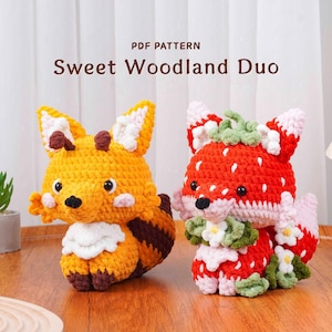 May include: Two crocheted fox figures. One is yellow and brown, the other is red and white, resembling a strawberry. The text "Sweet Woodland Duo" is above them. The figures are handmade and whimsical.
