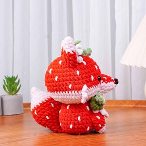 May include: A handmade crochet fox toy in a strawberry design. The fox is primarily red with white seed-like details, a white and pink trim, and green leaves. The fox has a black nose and is sitting on a wooden surface.