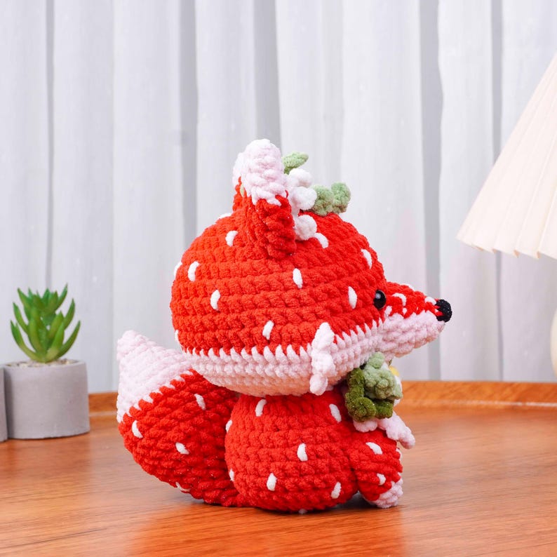 May include: A handmade crochet fox toy in a strawberry design. The fox is primarily red with white seed-like details, a white and pink trim, and green leaves. The fox has a black nose and is sitting on a wooden surface.