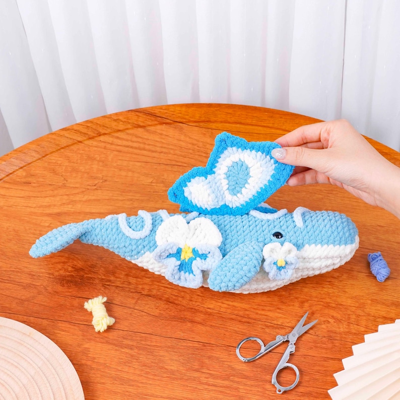 May include: A handcrafted crochet whale in blue and white, decorated with floral details. The whale's fin is held up, displaying its detailed design. Small scissors and yarn remnants are also visible.