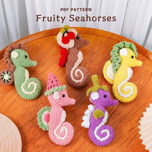 Puede incluir: Una colección de cinco caballitos de mar de ganchillo coloridos, cada uno diseñado para parecerse a una fruta diferente. Los caballitos de mar están dispuestos sobre una superficie de madera. El texto "PDF PATTERN Fruity Seahorses" está en la parte superior de la imagen.