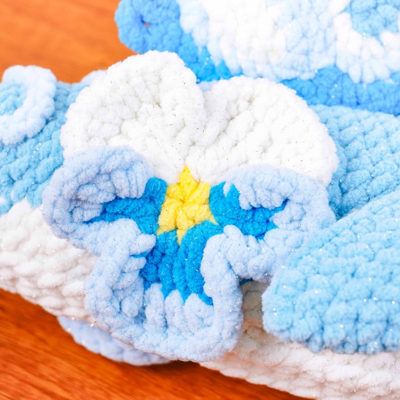 May include: Close-up of a handmade, plush floral design in shades of blue, white, and yellow. The flower has a textured, fluffy appearance, with a bright yellow centre and detailed petal work. The background is a wooden surface.
