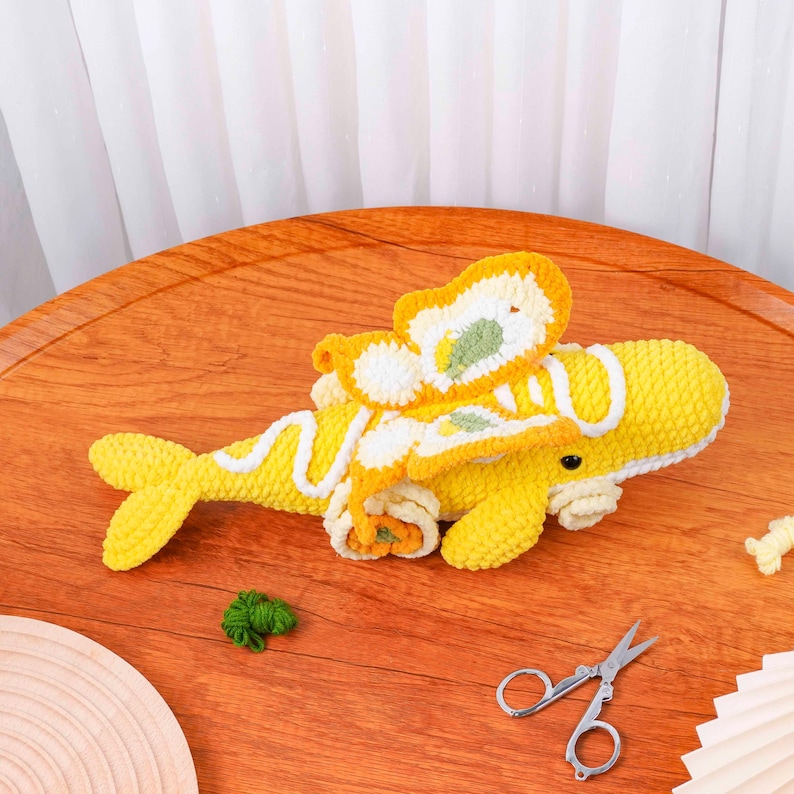 May include: A handmade yellow crochet whale with white detailing and a yellow and white crochet butterfly. The whale has a small black eye and a white belly. A pair of small scissors and a green yarn ball are on the wooden surface.