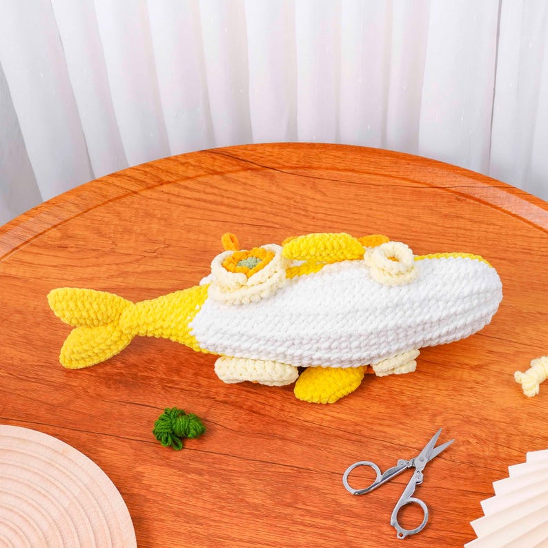 May include: A handcrafted crochet fish toy in yellow and white. The fish features a yellow tail, fins, and details, with a white body. Decorative elements include a flower-like design on top. The toy is placed on a wooden surface.