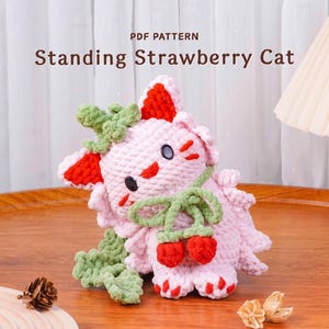 May include: A handmade pink crochet cat with red and green accents, resembling a strawberry. The cat has black eyes and is adorned with green leaves and red strawberry details. The text "PDF PATTERN Standing Strawberry Cat" is visible.