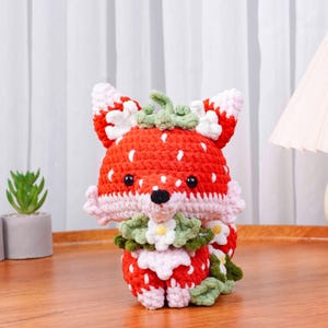 May include: A handmade crochet fox doll with a strawberry design. The fox is primarily red with white speckles, resembling a strawberry. It has pink and white accents, black eyes, and a green stem. The fox is sitting on a wooden surface.