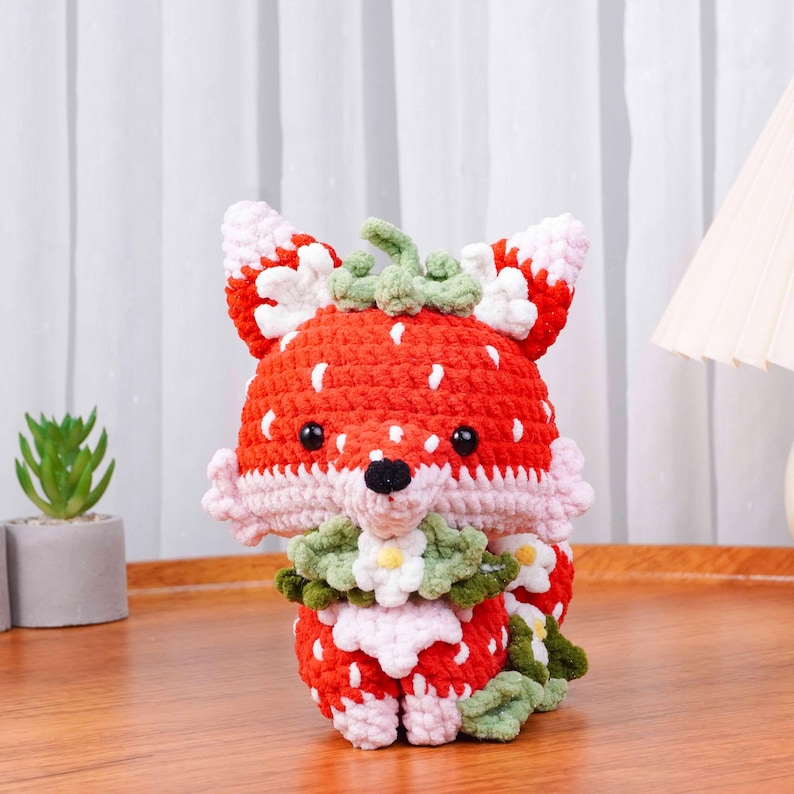 May include: A handmade crochet fox doll with a strawberry design. The fox is primarily red with white speckles, resembling a strawberry. It has pink and white accents, black eyes, and a green stem. The fox is sitting on a wooden surface.