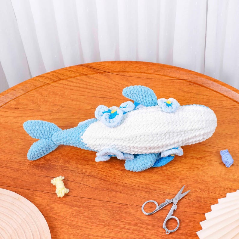May include: A handmade, crocheted whale plushie in shades of blue and white, with small flower accents. The whale has a detailed tail and fins, on a wooden surface. Small scissors and yarn scraps are also visible.