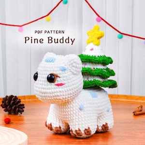 May include: A crocheted white creature with a Christmas tree on its back, topped with a yellow star. The creature has large black eyes, blue accents, and brown paws. The text "PDF PATTERN Pine Buddy" is visible.