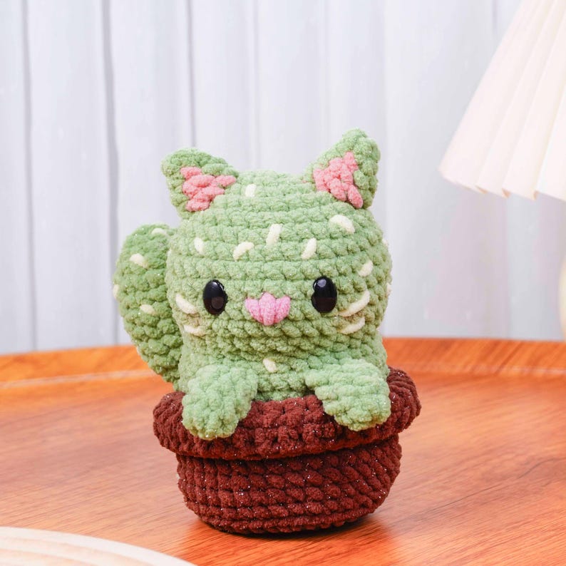 Op de afbeelding: Een handgemaakte gehaakte cactuskat in een bruine pot. De kat is groen met witte en roze accenten, waaronder roze oren en een roze neus. De pot is donkerbruin van kleur. De kat heeft zwarte ogen.