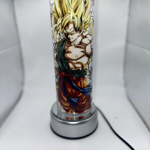 May include: A silver lamp base with a clear glass cylinder featuring a colorful anime character with yellow hair. The character is wearing an orange and red outfit with a blue belt.