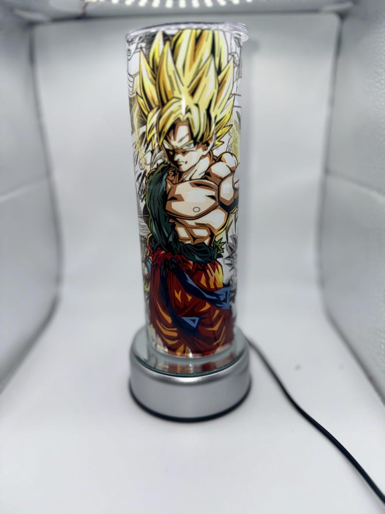 May include: A silver lamp base with a clear glass cylinder featuring a colorful anime character with yellow hair. The character is wearing an orange and red outfit with a blue belt.